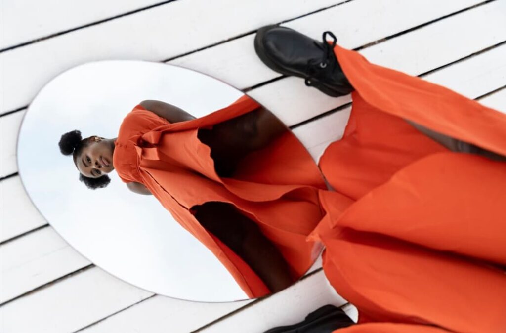 A woman in a flowing orange dress looks up, framed by a circular mirror set on a white wooden floor