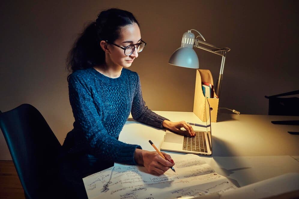 Woman working on a laptop and writing on paper in a dimly lit room with a table lamp
